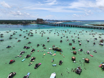 Aerial view of a turquoise shallow sandbar and coastal bay filled with dozens of anchored party boats, floating platforms and tiki huts, people wading in clear water, and a long causeway bridge to a barrier island under a sunny blue sky