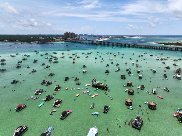 Aerial view of a turquoise shallow sandbar and coastal bay filled with dozens of anchored party boats, floating platforms and tiki huts, people wading in clear water, and a long causeway bridge to a barrier island under a sunny blue sky
