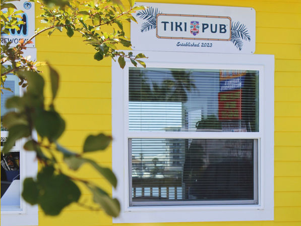 Bright yellow exterior of a tiki-themed pub with a white-framed window and closed blinds, leafy branch in the foreground and palm-tree reflections in the glass, coastal storefront vibe.