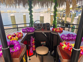 Tiki-roofed boat helm decked with bright purple and red flower leis, small steering wheel and stool overlooking a calm marina with wooden pilings and waterfront buildings.