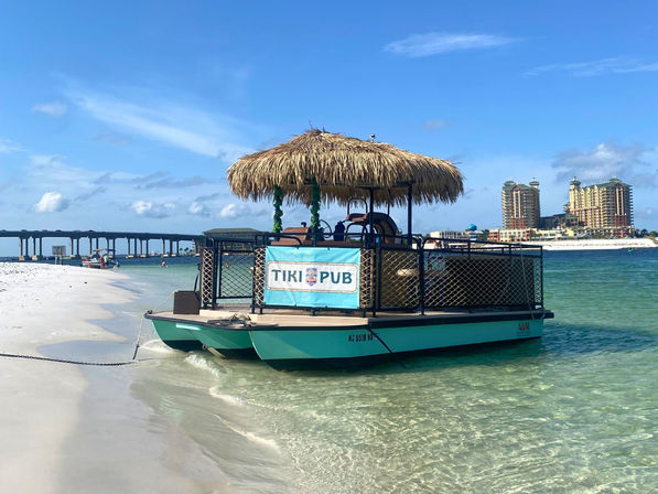 Thatched-roof tiki-style floating bar moored on a white-sand beach with crystal-clear turquoise water, a long seaside bridge to the left and beachfront high-rise condos on the horizon under a bright blue sky.
