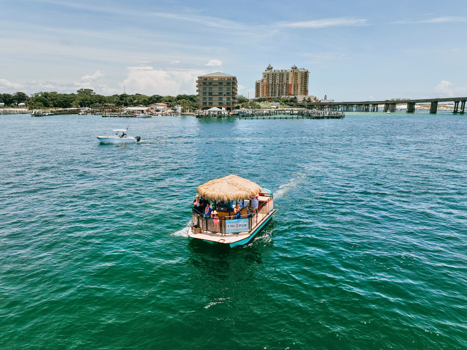 Thatched-roof tiki pontoon with passengers cruising turquoise bay toward a coastal marina, framed by a small powerboat, beachfront resort buildings and a long bridge