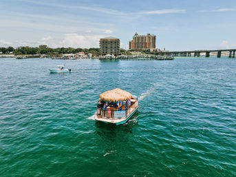 Thatched-roof tiki pontoon with passengers cruising turquoise bay toward a coastal marina, framed by a small powerboat, beachfront resort buildings and a long bridge