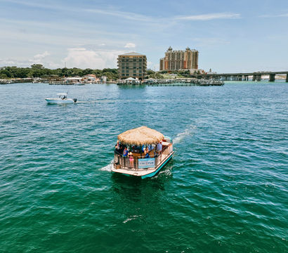 Thatched-roof tiki pontoon with passengers cruising turquoise bay toward a coastal marina, framed by a small powerboat, beachfront resort buildings and a long bridge