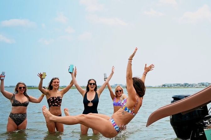 Five friends in swimsuits cheer in shallow water by a boat as one slides off a boat slide into the bay, others raising drinks and arms against a sunny shoreline.
