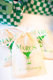 Cream drawstring favor bags printed with a green margarita cocktail graphic and script text, arranged beside a green-and-white checkered pouch — fun margarita-themed wedding favors
