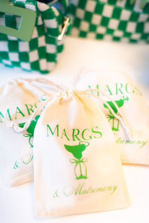 Cream drawstring favor bags printed with a green margarita cocktail graphic and script text, arranged beside a green-and-white checkered pouch — fun margarita-themed wedding favors