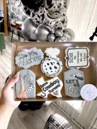 Hand holding a box of bridal bachelorette cookies in silver, gray and cow-print: diamond ring cookie, Texas-shaped “Same Cowboy Forever” cookie, disco leg and message tags, silver and black balloon garland in background.