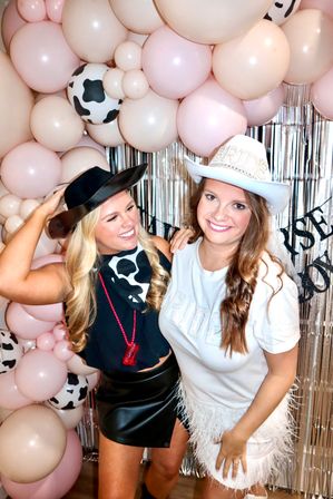 Two friends smiling in a cowgirl-themed bachelorette party photo booth, one wearing a white ‘BRIDE’ cowboy hat and feathered skirt and the other in a black hat with a cow-print bandana, posed in front of pink and cow-print balloon arch and metallic fringe backdrop.