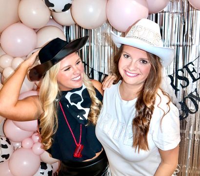 Two friends smiling in a cowgirl-themed bachelorette party photo booth, one wearing a white ‘BRIDE’ cowboy hat and feathered skirt and the other in a black hat with a cow-print bandana, posed in front of pink and cow-print balloon arch and metallic fringe backdrop.