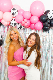 Two smiling women posing at an indoor party photo booth with a pink, black, and cow-print balloon arch and silver fringe backdrop.