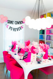 Bright modern dining room styled for a pink party with fuchsia velvet chairs around a white table topped with hot-pink gift bags, tissue paper, candy-filled goblets, scattered confetti, pink balloons and a black-letter banner reading "SAME COWBOY FO REVER", beneath a cloud-like pendant light and colorful bookshelves.