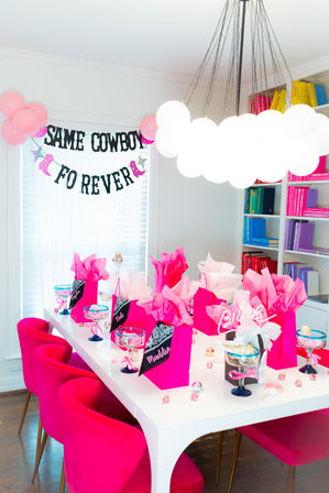 Bright modern dining room styled for a pink party with fuchsia velvet chairs around a white table topped with hot-pink gift bags, tissue paper, candy-filled goblets, scattered confetti, pink balloons and a black-letter banner reading "SAME COWBOY FO REVER", beneath a cloud-like pendant light and colorful bookshelves.