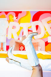 Playful white embroidered knee-high cowboy boots with wooden heels propped on a bed in a colorful retro bedroom with bold pink, yellow and red mural and oversized white balloon letters.