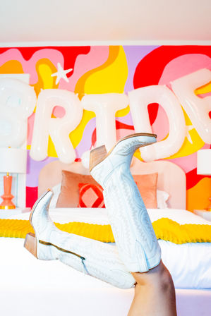 Playful white embroidered knee-high cowboy boots with wooden heels propped on a bed in a colorful retro bedroom with bold pink, yellow and red mural and oversized white balloon letters.