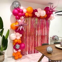 Vibrant party backdrop with pink, fuchsia and orange balloon garland, metallic star and disco-ball accents framing a pink foil fringe curtain in a modern dining room setting.
