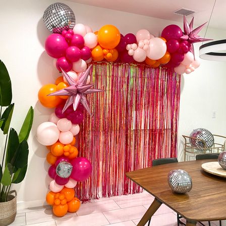 Vibrant party backdrop with pink, fuchsia and orange balloon garland, metallic star and disco-ball accents framing a pink foil fringe curtain in a modern dining room setting.