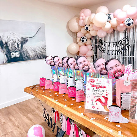 Cowboy-themed bachelorette party setup with pink and cream balloon garland and cow-print balloons, silver fringe backdrop with playful banner, wooden table of holographic favor bags topped with repeated male face cutouts, welcome sign and cowboy boot decorations.