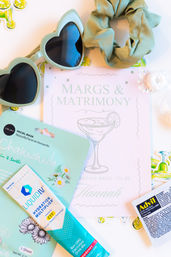Mint-green bachelorette flat lay: heart-shaped sunglasses, scrunchie, chamomile facial sheet mask, hydration drink sachet, pain reliever packet, and a 'Margs & Matrimony' bride‑to‑be card.