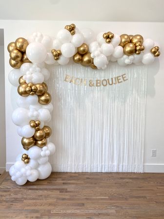 White and gold balloon garland framing a white fringe photo backdrop on hardwood floor, featuring gold 'BACH & BOUJEE' letters — elegant bachelorette party decor.