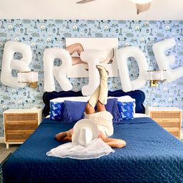 Playful bridal bedroom scene with large white "BRIDE" balloons above a navy quilted bed; person lies on bed with legs up in cream cowboy boots, holding a white wedding hat and veil, set against blue patterned wallpaper and rattan nightstands.