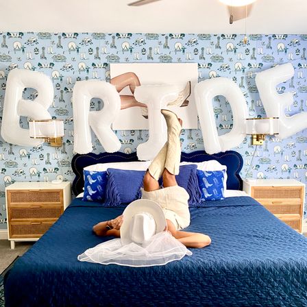 Playful bridal bedroom scene with large white "BRIDE" balloons above a navy quilted bed; person lies on bed with legs up in cream cowboy boots, holding a white wedding hat and veil, set against blue patterned wallpaper and rattan nightstands.