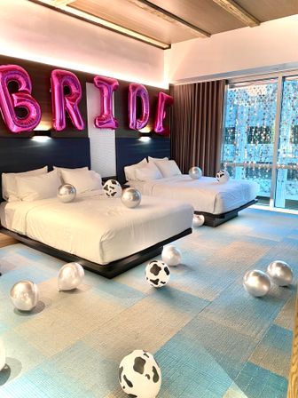 Bride-themed hotel room with two white-sheet beds, large pink foil 'BRIDE' balloons on the headboard, silver and cow-print balloons scattered on blue-check carpet, and a floor-to-ceiling window with star string lights and balcony view.