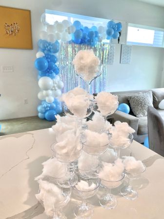 Living room baby shower decor with blue-and-white balloon arch, iridescent backdrop, and a champagne glass tower filled with fluffy cotton candy