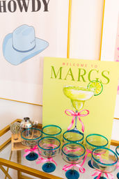 Colorful margarita bar cart with lime-green "Welcome to Margs" sign, illustrated margarita with lime, turquoise-rimmed glasses tied with pink ribbons, stainless cocktail shaker and bar tools on a gold serving cart