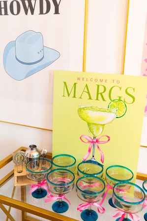 Colorful margarita bar cart with lime-green "Welcome to Margs" sign, illustrated margarita with lime, turquoise-rimmed glasses tied with pink ribbons, stainless cocktail shaker and bar tools on a gold serving cart