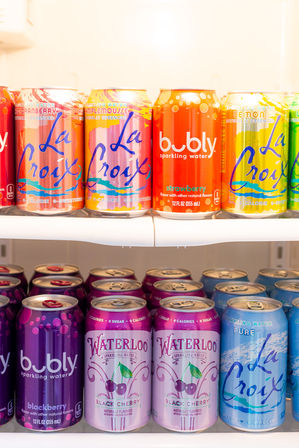 Bright, colorful sparkling water cans (La Croix, bubly, Waterloo) neatly lined on refrigerator shelves, showing lemon, strawberry and black cherry flavors.