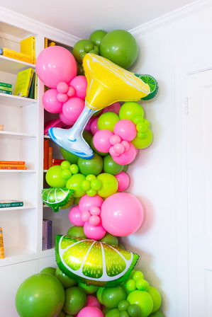 Playful pink and lime-green balloon garland in an indoor corner by a white bookshelf and door, featuring a yellow cocktail-glass foil balloon and lime-slice foil accents — festive party decor