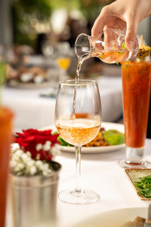 Hand pouring sparkling rosé into a wine glass on a sunlit outdoor patio table set for brunch with a Bloody Mary, plates, and a red flower centerpiece.