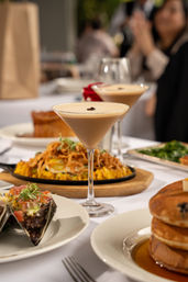 Restaurant brunch spread with creamy espresso martinis garnished with coffee beans, stacked pancakes with syrup, savory plated appetizers and a skillet of crispy rice on a white tablecloth, blurred diners in the background