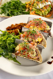 Three brunch tacos with shredded beef, scrambled eggs, pico de gallo, pickled onions and microgreens in a metal taco rack, served with arugula salad and seasoned roasted potatoes on a white plate