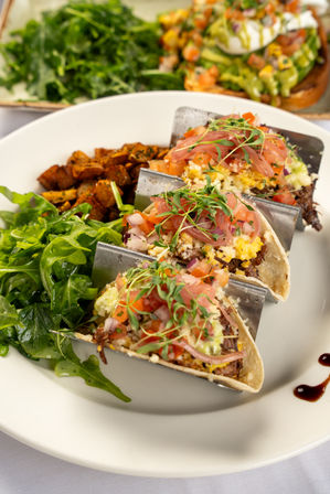Three brunch tacos with shredded beef, scrambled eggs, pico de gallo, pickled onions and microgreens in a metal taco rack, served with arugula salad and seasoned roasted potatoes on a white plate