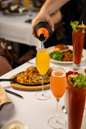 Hand pouring champagne into a bright orange mimosa on a white-tablecloth brunch table with skillet hash, a burger and Bloody Marys.