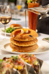 Close-up of a stack of three golden blueberry pancakes on a white plate with a pat of butter and maple syrup being poured, restaurant brunch setting with blurred wine glass and cocktail in the background