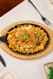 Cast-iron skillet on a wooden board with cheesy corn hash topped with crispy fried onion strings, microgreens and creamy sauce on a white restaurant table