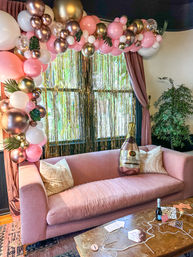 Celebration photo area in an indoor lounge: pink velvet sofa under a pink, white and rose-gold balloon garland with tropical leaf accents, metallic gold fringe backdrop, oversized champagne balloon and party props on a coffee table.