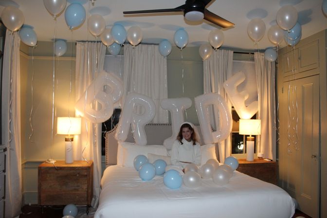 Cozy bedroom decked out for a bridal celebration with large letter balloons spelling BRIDE, pale blue and ivory balloons floating from the ceiling and scattered on the bed where a woman in white sits between glowing bedside lamps.