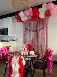 Festive cherry-themed party setup in a modern apartment kitchen dining area: pink, red and white balloon garland with green cherry accents above a metallic pink fringe backdrop, round glass table with stacked pink cups and a thank-you card, and bright feather boas draped over black chairs.