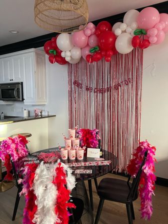 Festive cherry-themed party setup in a modern apartment kitchen dining area: pink, red and white balloon garland with green cherry accents above a metallic pink fringe backdrop, round glass table with stacked pink cups and a thank-you card, and bright feather boas draped over black chairs.