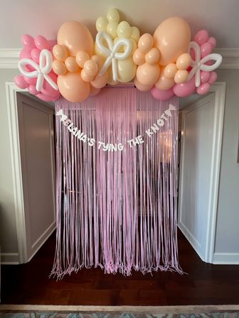 Pastel balloon arch of peach, pink and pale yellow balloons with white balloon bow accents over a doorway, pink fringe curtain hanging below and a white letter banner — festive wedding-shower photo backdrop.