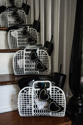 White plastic mesh baskets lined up on indoor wooden stairs, each containing black rhinestone-trimmed heart sunglasses, a black travel tumbler with straw and folded black apparel — playful party-favor display.