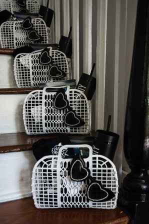 White plastic mesh baskets lined up on indoor wooden stairs, each containing black rhinestone-trimmed heart sunglasses, a black travel tumbler with straw and folded black apparel — playful party-favor display.