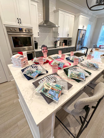 Modern white kitchen with marble island and stainless appliances, island arranged with multiple gift bags of snacks, folded shirts and pink party cups for a casual gathering.