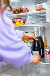 Person’s hands reaching into a bright home refrigerator to grab a chilled bottle of sparkling wine, with fresh strawberries, a mixed fruit bowl and canned drinks on the shelves.