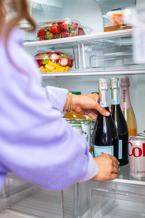 Person’s hands reaching into a bright home refrigerator to grab a chilled bottle of sparkling wine, with fresh strawberries, a mixed fruit bowl and canned drinks on the shelves.