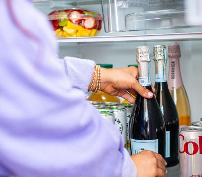 Person’s hands reaching into a bright home refrigerator to grab a chilled bottle of sparkling wine, with fresh strawberries, a mixed fruit bowl and canned drinks on the shelves.